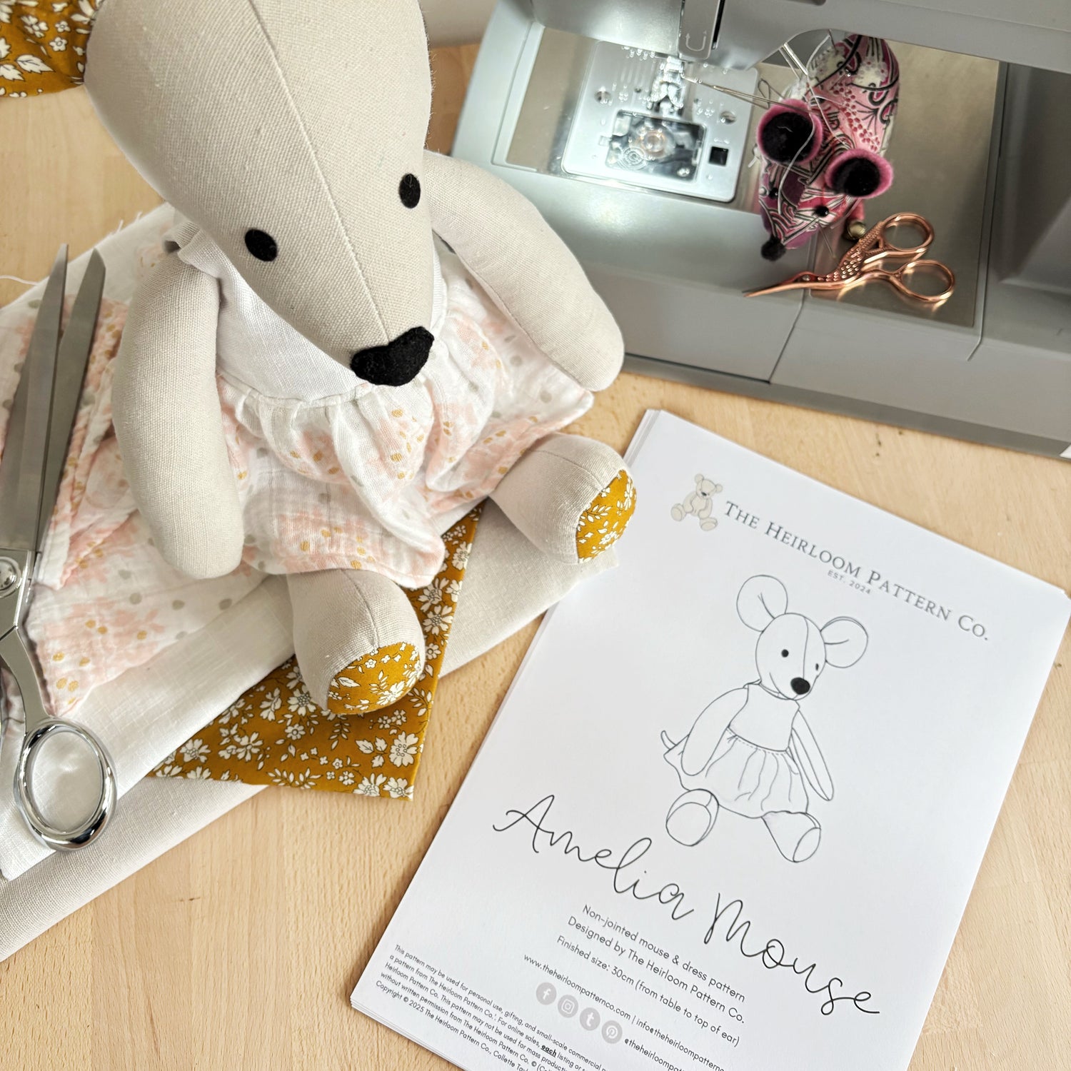 Stuffed Amelia mouse toy, sewing machine, scissors, and pattern sheet on a wooden surface with The Heirloom Pattern Co branding.